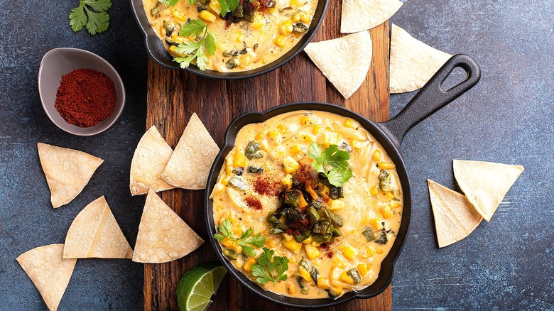 Hot corn dip in two small cast iron skillets surrounded by corn chips on a wood serving board