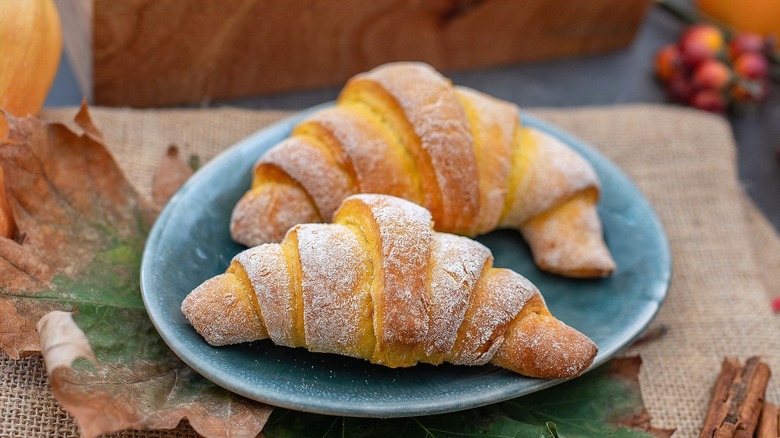 Two baked crescent rolls with powdered sugar on a blue plate surrounded by fall leaves and cinnamon sticks