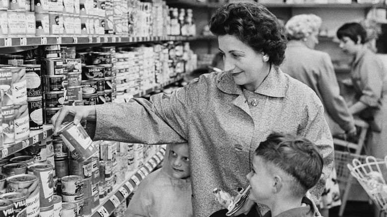 A black and white image of a woman and two children shopping for canned goods in the 1960s