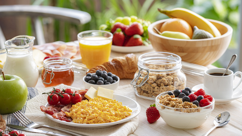 Table filled with assorted breakfast staples including yogurt, scrambled eggs, milk, juice, granola, and fruits