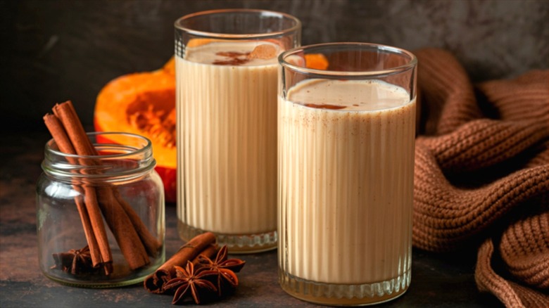 Two pumpkin smoothies in tall clear glasses next to a jar of cinnamon sticks and star anise pods