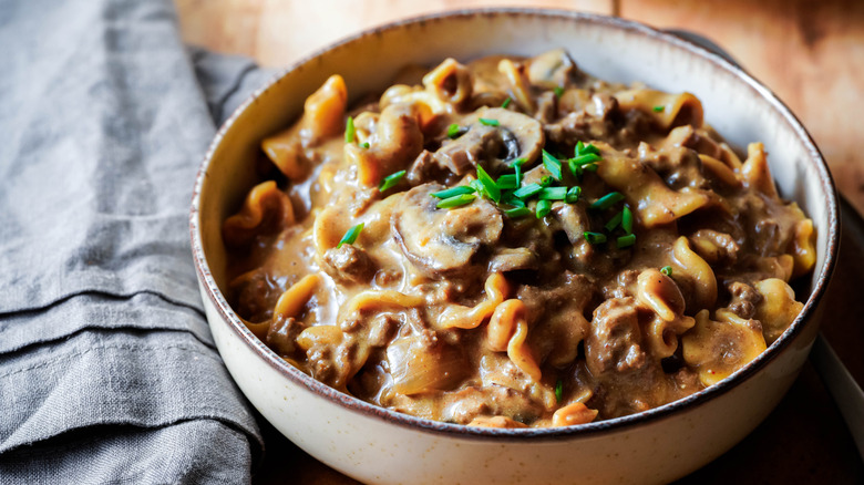 A bowl of fresh ground beef stroganoff served over egg noodles and topped with chopped mushrooms and diced herbs