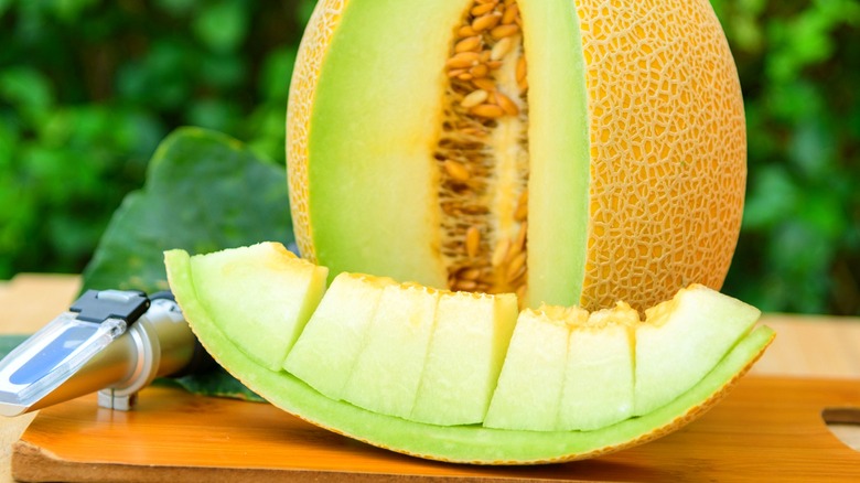 Part of a Galia melon sliced into chunks in front of the rest of the melon on a wooden cutting board