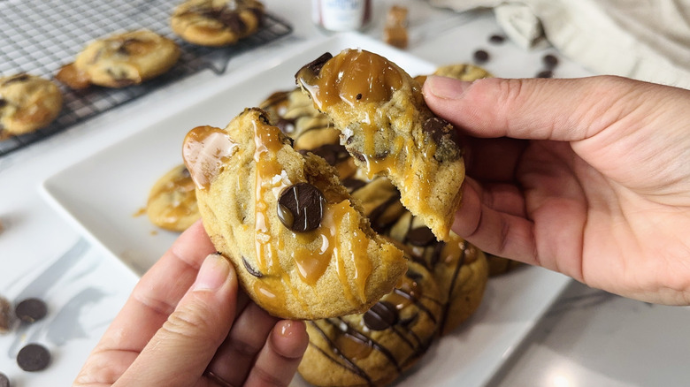 hands breaking chocolate caramel cookies in half with plate of cookies in the background