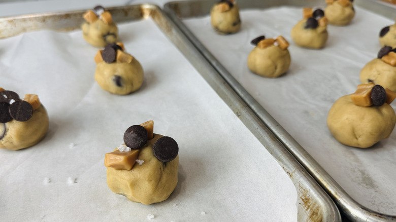 cookie dough balls topped with chocolate chips, salt, and caramel on lined baking trays