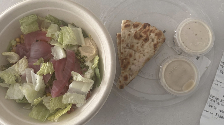 Salad in a container next to a lid with pita bread and two sauce containers
