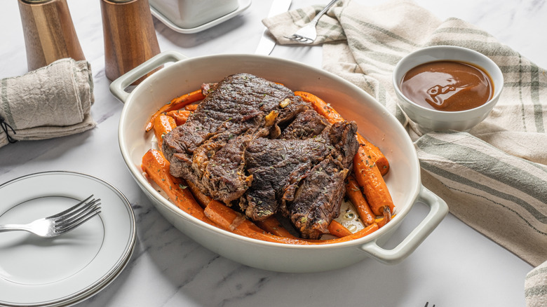 A standard pot roast with carrots in a white dish on a table surrounded by various table settings.