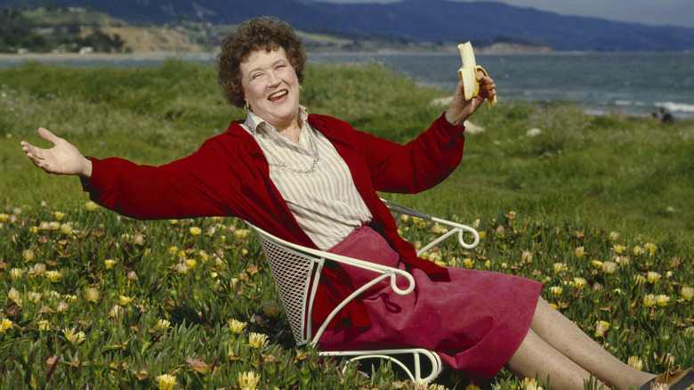 Julia Child smiling, eating a banana in a coastal field of flowers