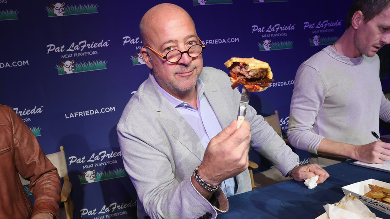 Andrew Zimmern holding a hamburger on a knife at a cooking competition