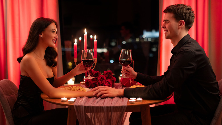 Couple holding hands and drinking wine at a restaurant with candlelight and red roses on the table.