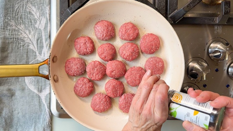 Seasoning small beef patties in large white skillet with pepper grinder
