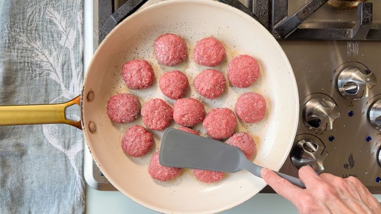 Pressing down on small beef patties cooking in large white skillet with spatula