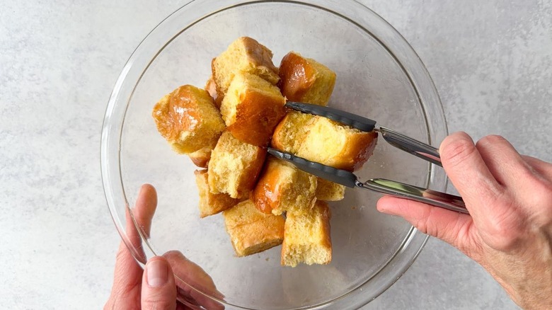 Tossing brioche cubes with melted butter in glass bowl using tongs