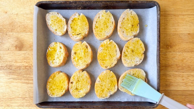 Brushing olive oil and minced garlic on baguette slices on parchment-lined baking sheet with pastry brush