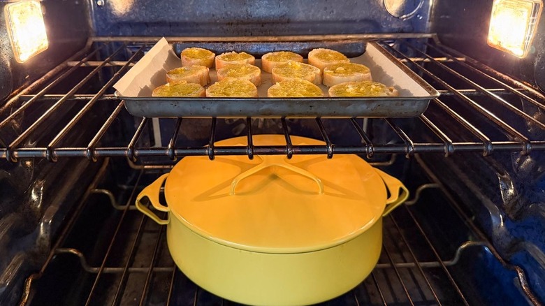 Tray of baguette croutons baking above covered yellow braiser in oven
