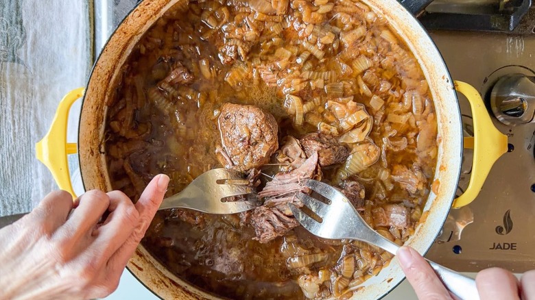 Shredding French onion pot roast into chunks with forks in large pot on stovetop