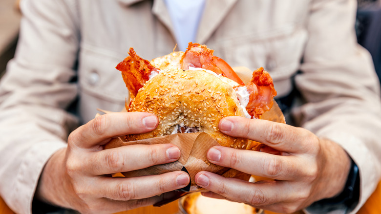 A man holds a breakfast bagel with cream cheese and bacon