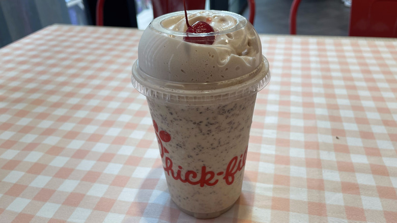 A plastic cup with a cherry-topped S'mores Milkshake from Chick-fil-A with chocolate bits sits on a checkered tablecloth.