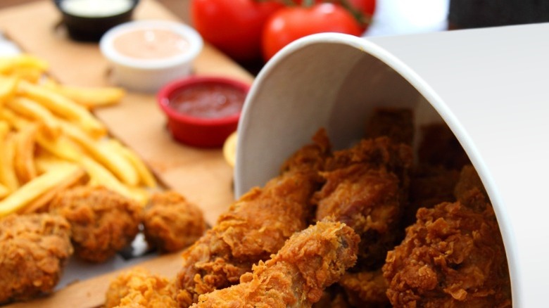 A bucket of fast food chicken pouring out onto a table with fries behind it.