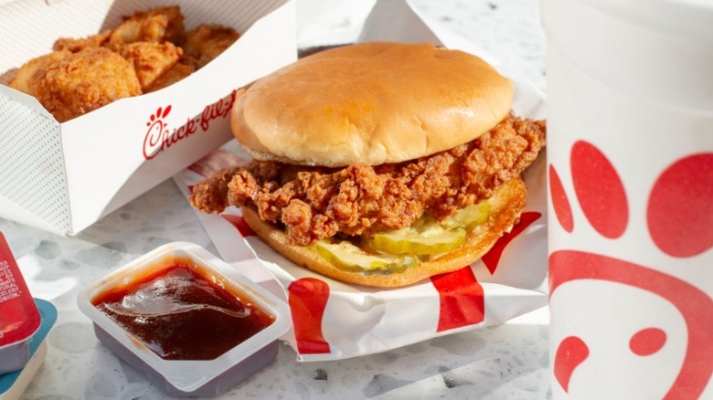 A Chick-fil-A chicken sandwich and chicken bites with polynesian sauce siting on a table with additional sauce packets and a drink off to the side.