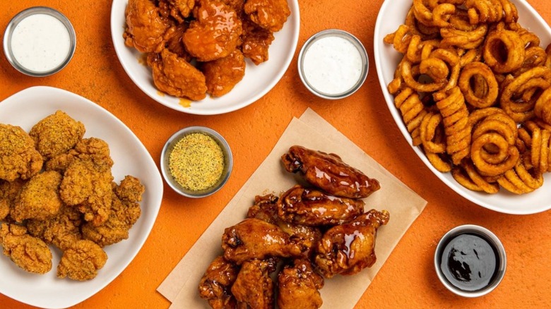 Variety of wings and curly fries from It's Just Wings with small bowls of dipping sauces.