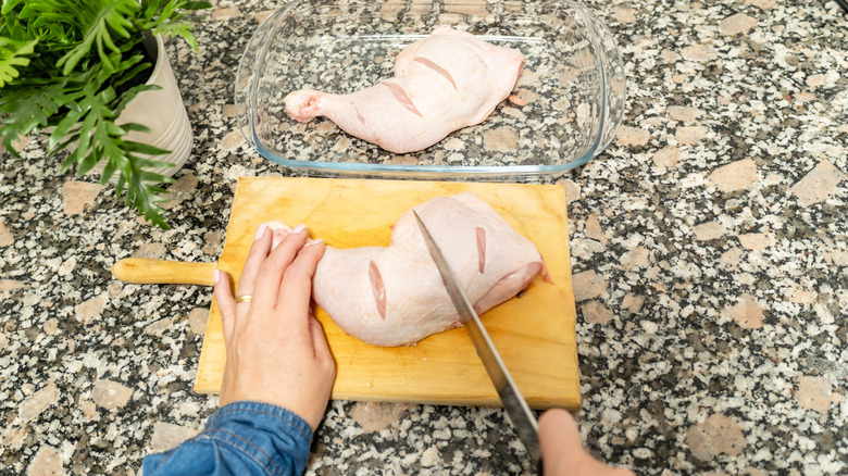 Woman scoring raw chicken thighs on cutting board with a raw chicken quarter in glass dish.