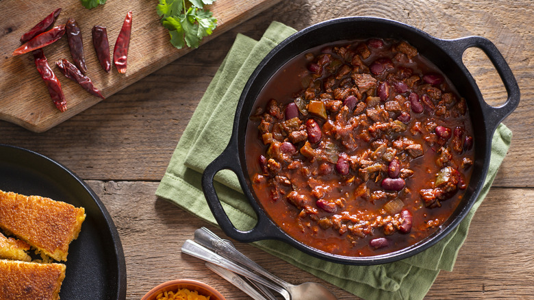 Chili in a cast-iron pot on a green cloth with cornbread, cheese, and chili peppers on a wooden table