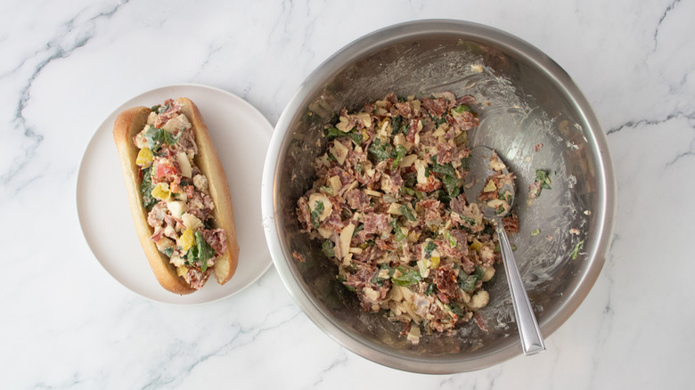 A filled roll next to a large bowl of chopped Italian sandwich ingredients