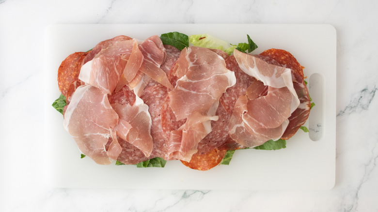 Assorted sliced Italian deli meats and chopped lettuce on cutting board