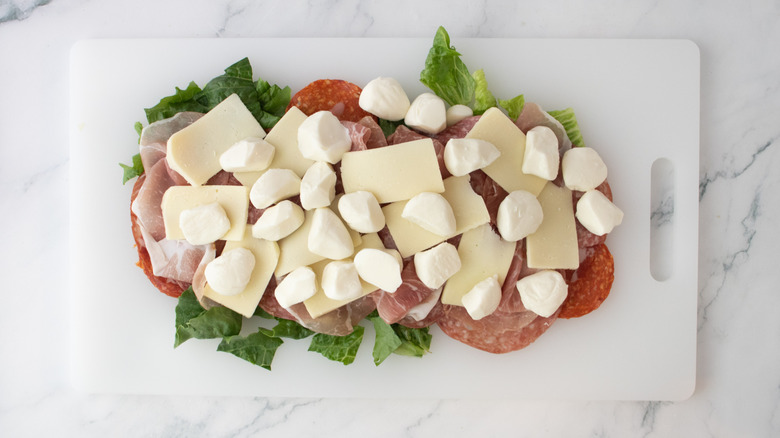 A mix of fresh and sliced cheese, Italian deli meats, and lettuce on cutting board