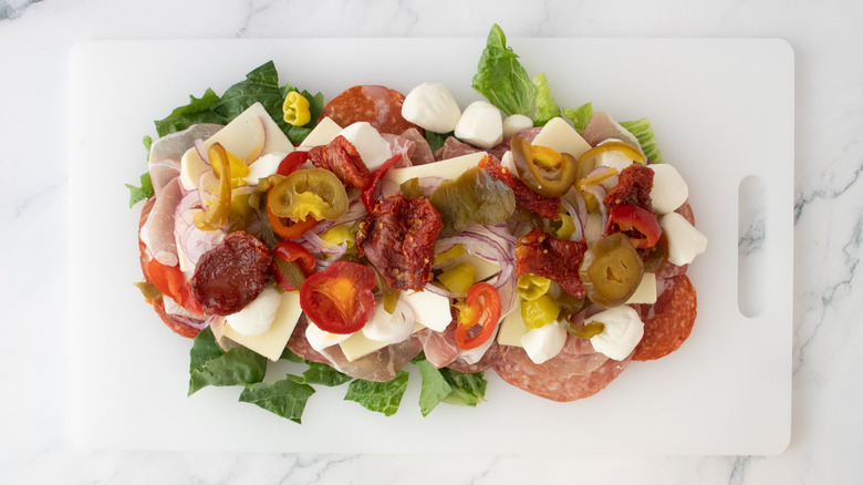 A mix of jarred vegetables, cheese, Italian deli meats, and lettuce on cutting board