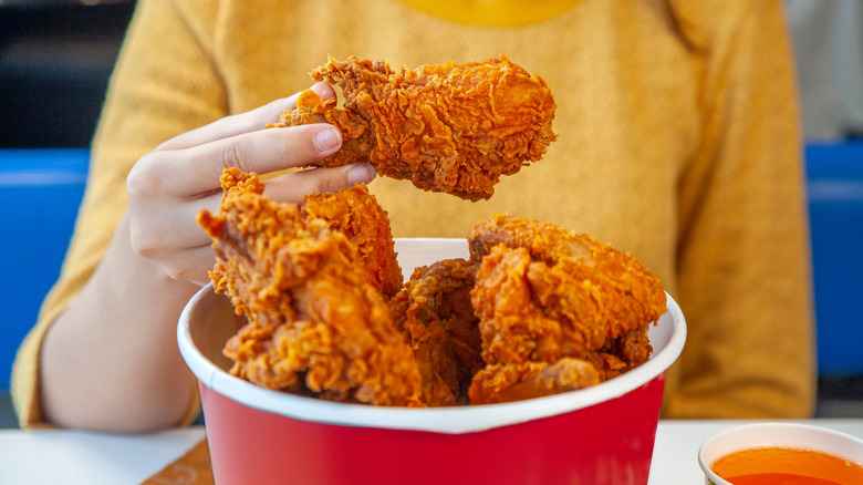 Person in a yellow shirt holding a piece of fried chicken over a bucket full of fried chicken