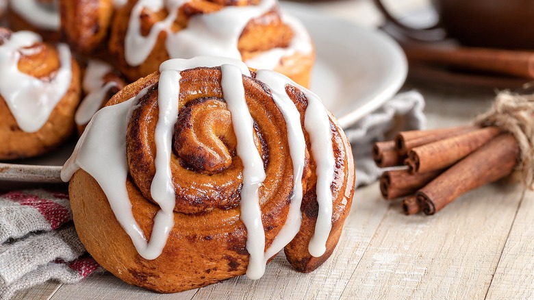 A cinnamon roll with white icing leaning on the edge of a plate next to a bundle of cinnamon sticks