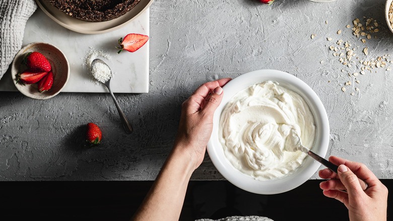 A person's hands are seen stirring whipped cream and a bowl; sliced strawberries are also in the frame.