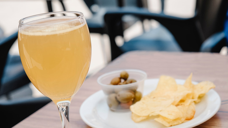A glass with shandy -- beer mixed with lemonade -- sits on a table next to a plate with some potato chips and some green olives, which is a popular appetizer in spain
