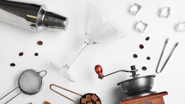 a martini glass, cocktail shaker, grinder, and sieve with coffee beans and ice cubes scattered around them