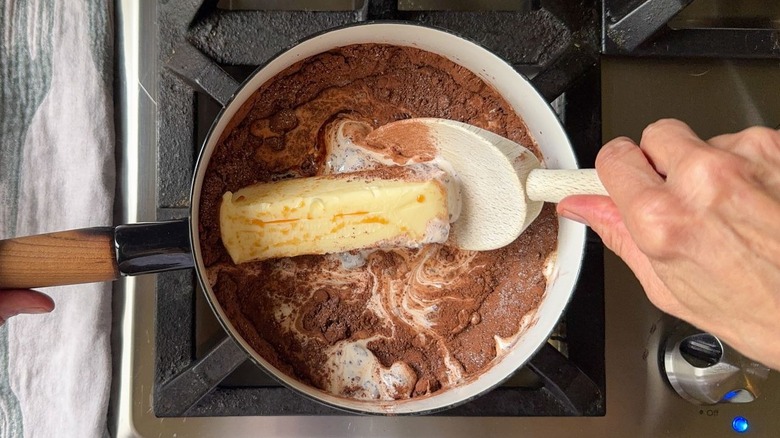 Butter, cream, cocoa powder, and sugar in saucepan on stovetop with wooden spoon