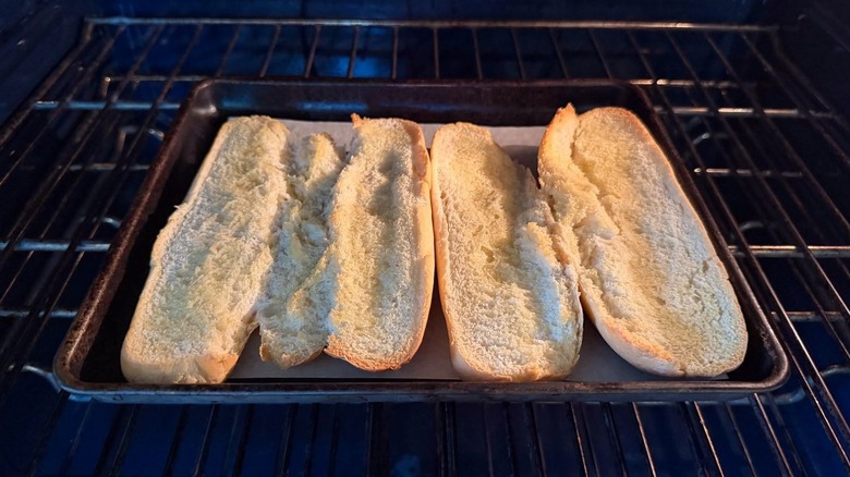Sliced Italian rolls with olive oil toasting on parchment-lined baking sheet