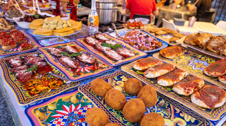 Spread of Sicilian street foods on colorful plates, including stuffed eggplant, arancini, bruschetta, and more