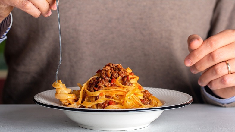 Hand twirling linguini noodles with meat sauce on a metal fork from a white bowl with black trim