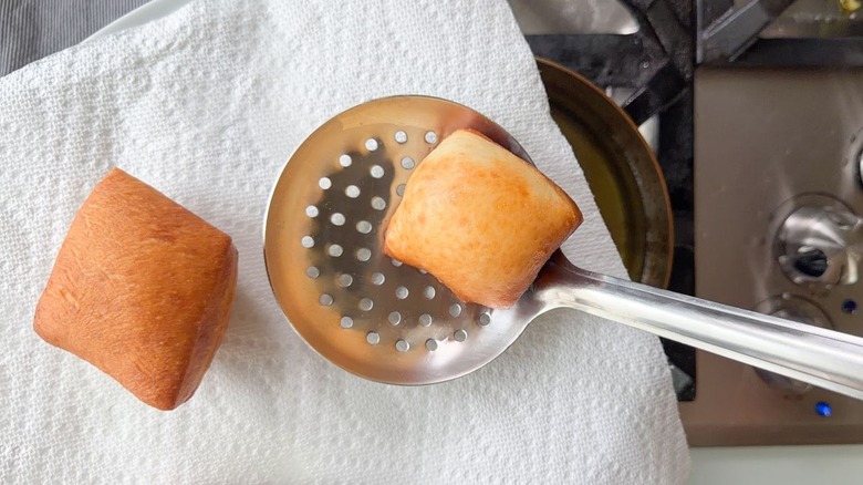 Using a slotted spoon to transfer hot beignets to paper towels on a plate