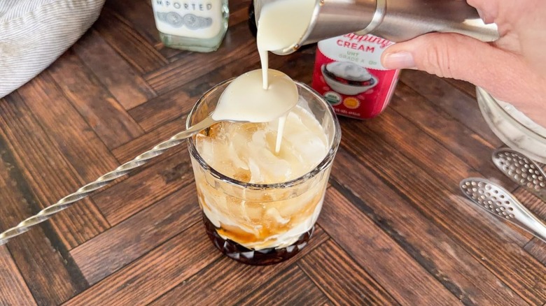 Pouring heavy cream over a spoon into a White Russian cocktail in rocks glass