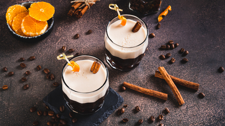 Two carajillo cocktails on a table with coffee beans and orange slices and cinnamon sticks