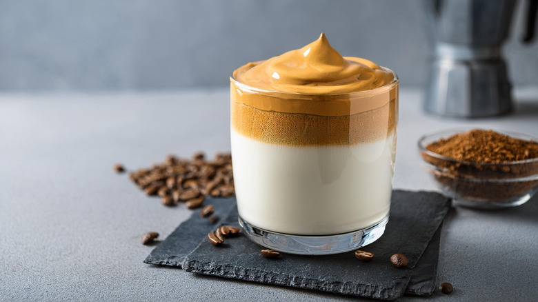 A dalgona coffee in a glass on a coaster in front of coffee beans and a dish of coffee grounds