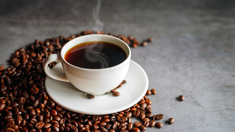 A steaming cup of hot coffee on a saucer, surrounded by roasted coffee beans.