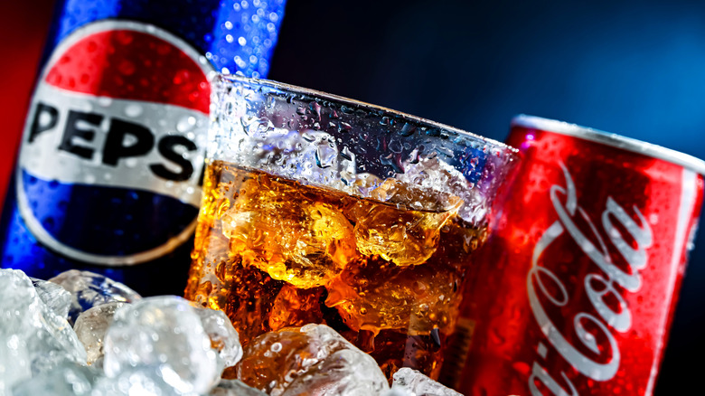 A can of Coke and a can of Pepsi each sitting on ice with one glass of unmarked soda and ice