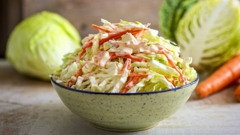 A speckled grey-blue stoneware bowl filled with a mound of creamy coleslaw set in front of cabbages and carrots
