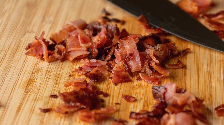 Chopped, fully-cooked bacon near the blade of a sharp knife on a wooden cutting board