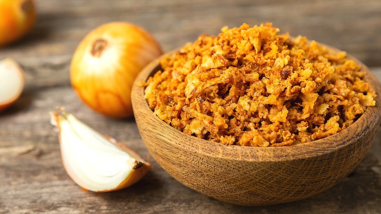 Crispy fried onions in a wooden bowl next to fresh, whole, and quartered yellow onions