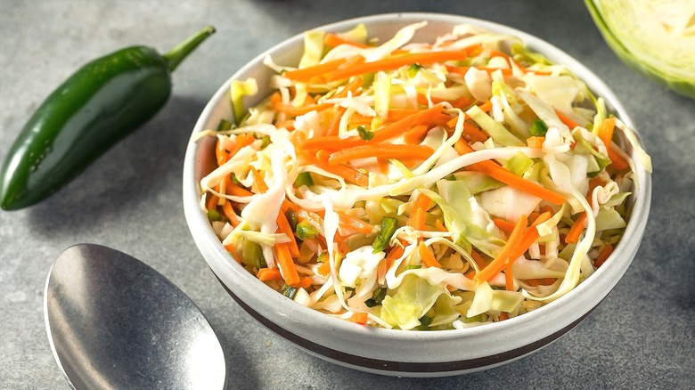 Bowl of coleslaw with sliced cabbage, shredded carrot, and chopped jalapeño on a gray surface